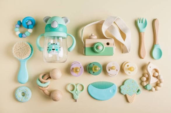 Lasten sinivihreitä ja luonnonvärisiä leluja ja vauvan tarvikkeita, kuten tuttia, ruokailuvälineitä, harjaa ja lelukameraa vaalealla taustalla.