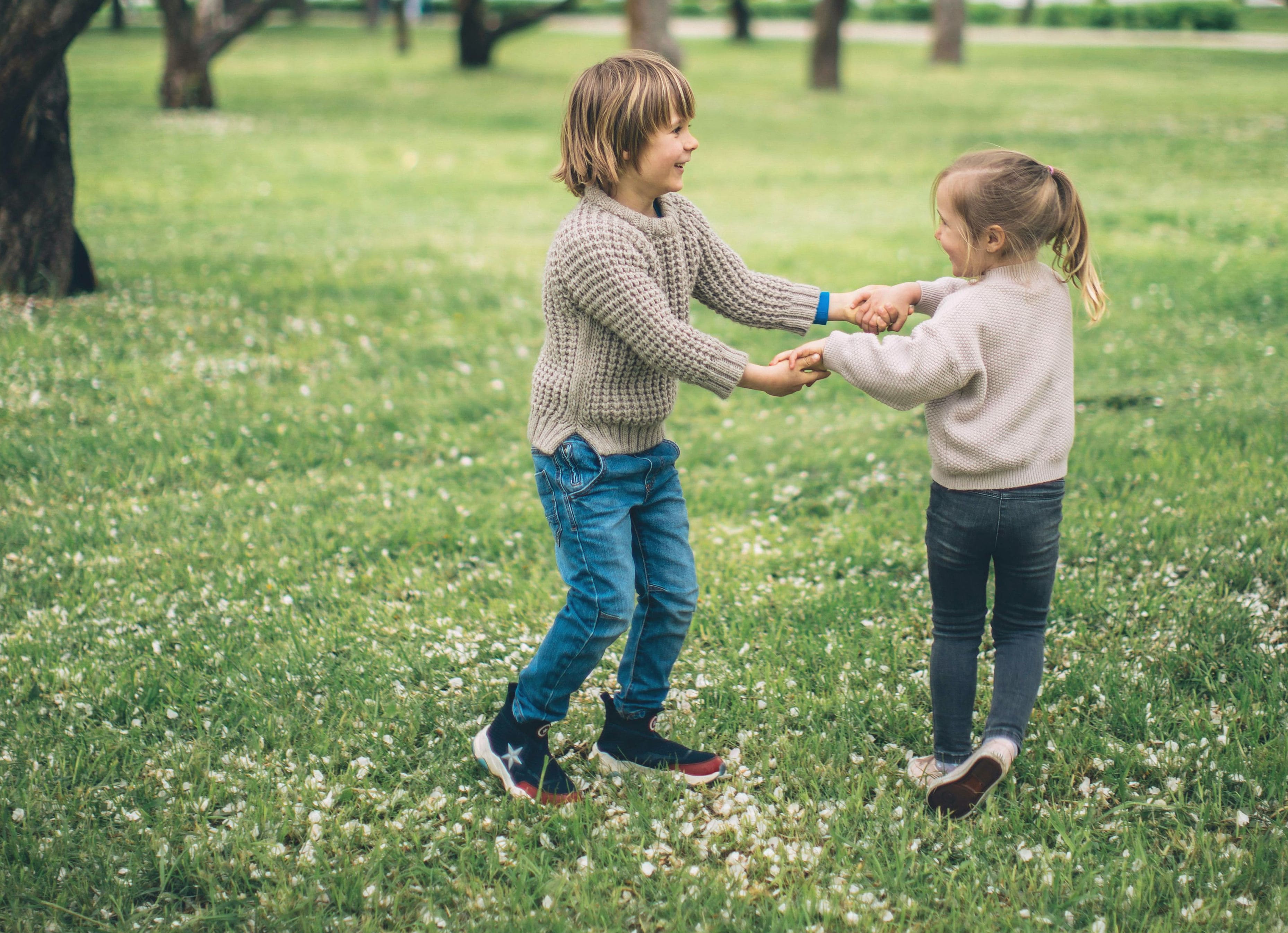 Kaksi lasta pitää toisiaan kädestä ja leikkii nurmikolla ulkona.
