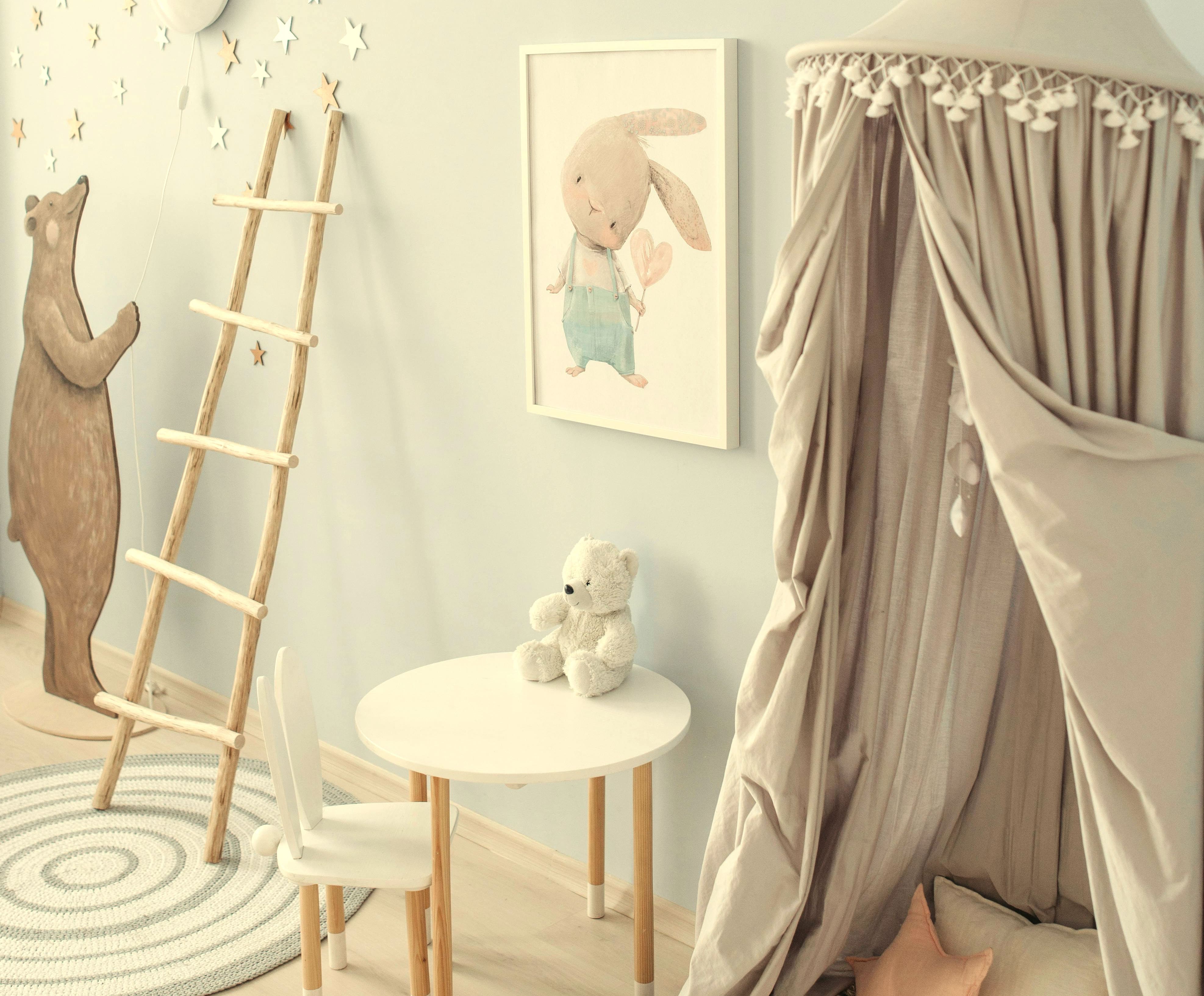 Lastenhuone, jossa valkoinen pöytä ja tuoli, nallepieni pöydällä, seinällä pupujuliste, puiset tikkaat ja kangasleikkiteltta tyynyillä.