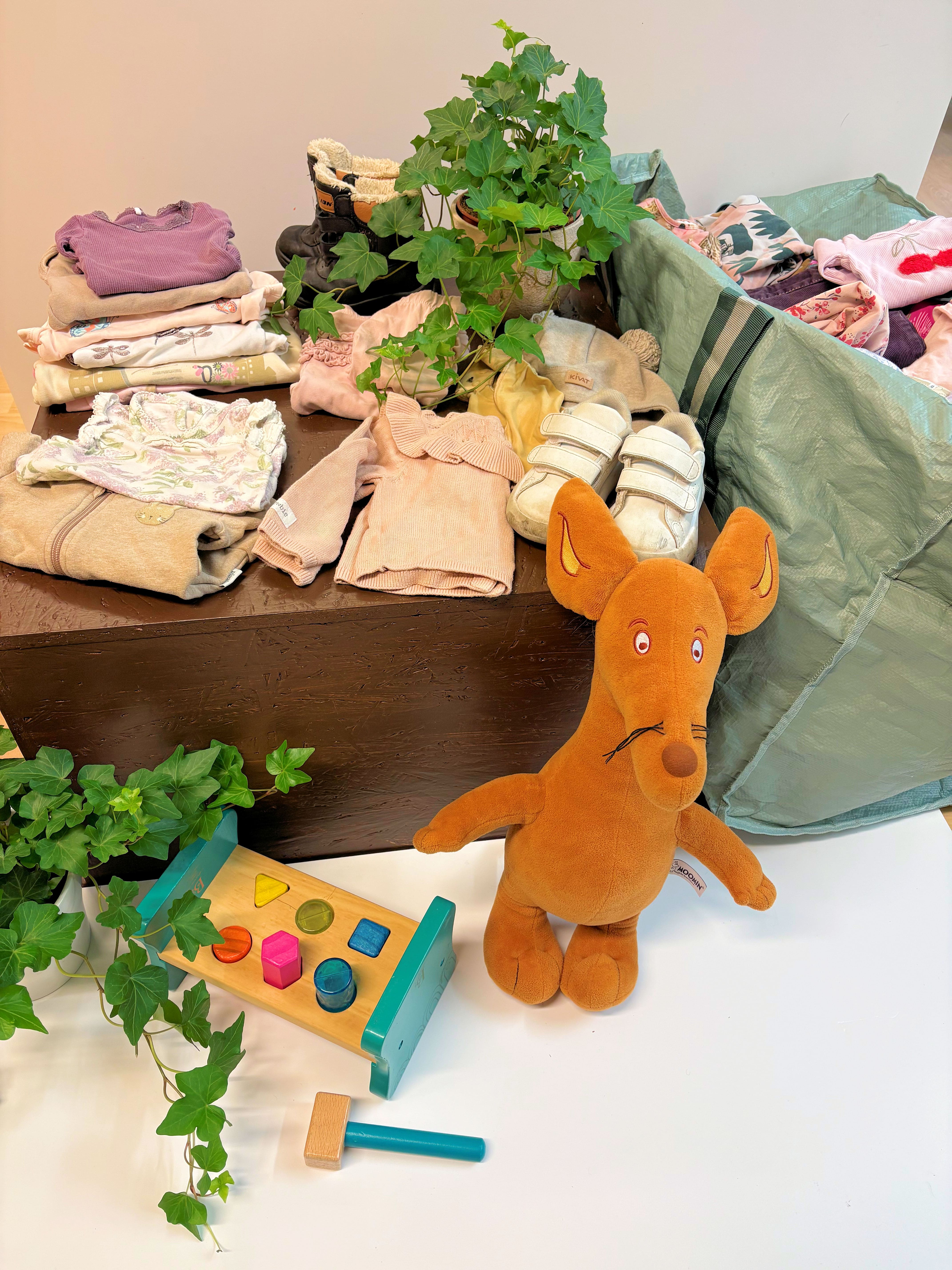 Lastenvaatteita, leluja ja pehmolelu ruskean pöydän päällä ja viereisessä vihreässä säilytyskorissa.