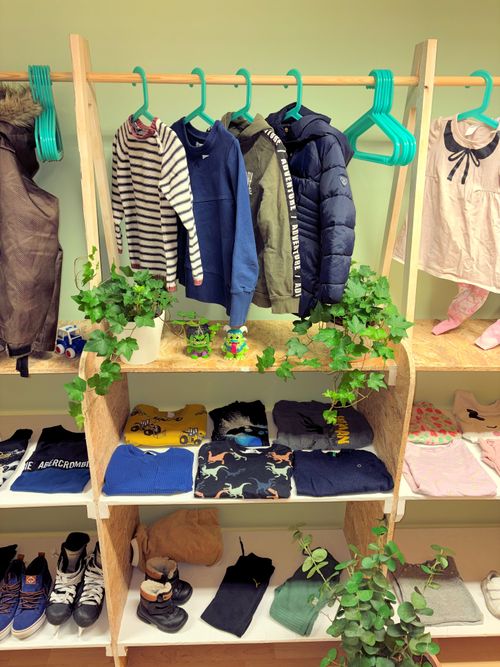 Lasten vaatteita ripustettuna ja taiteltuna puisessa hyllyssä, mukana takkeja, puseroita, housuja ja kenkiä, sekä koristekasveja.