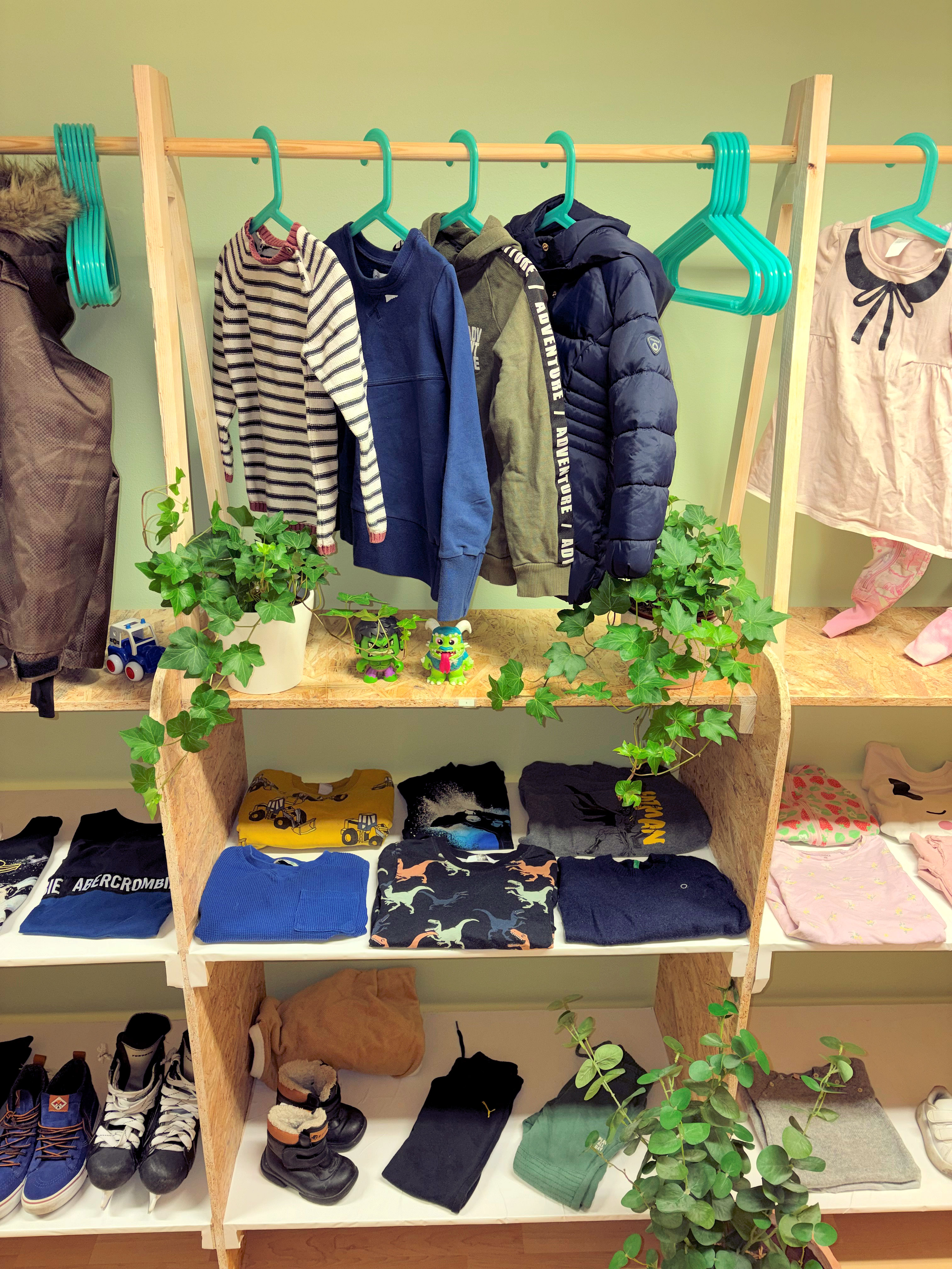Lasten vaatteita ripustettuna ja taiteltuna puisessa hyllyssä, mukana takkeja, puseroita, housuja ja kenkiä, sekä koristekasveja.