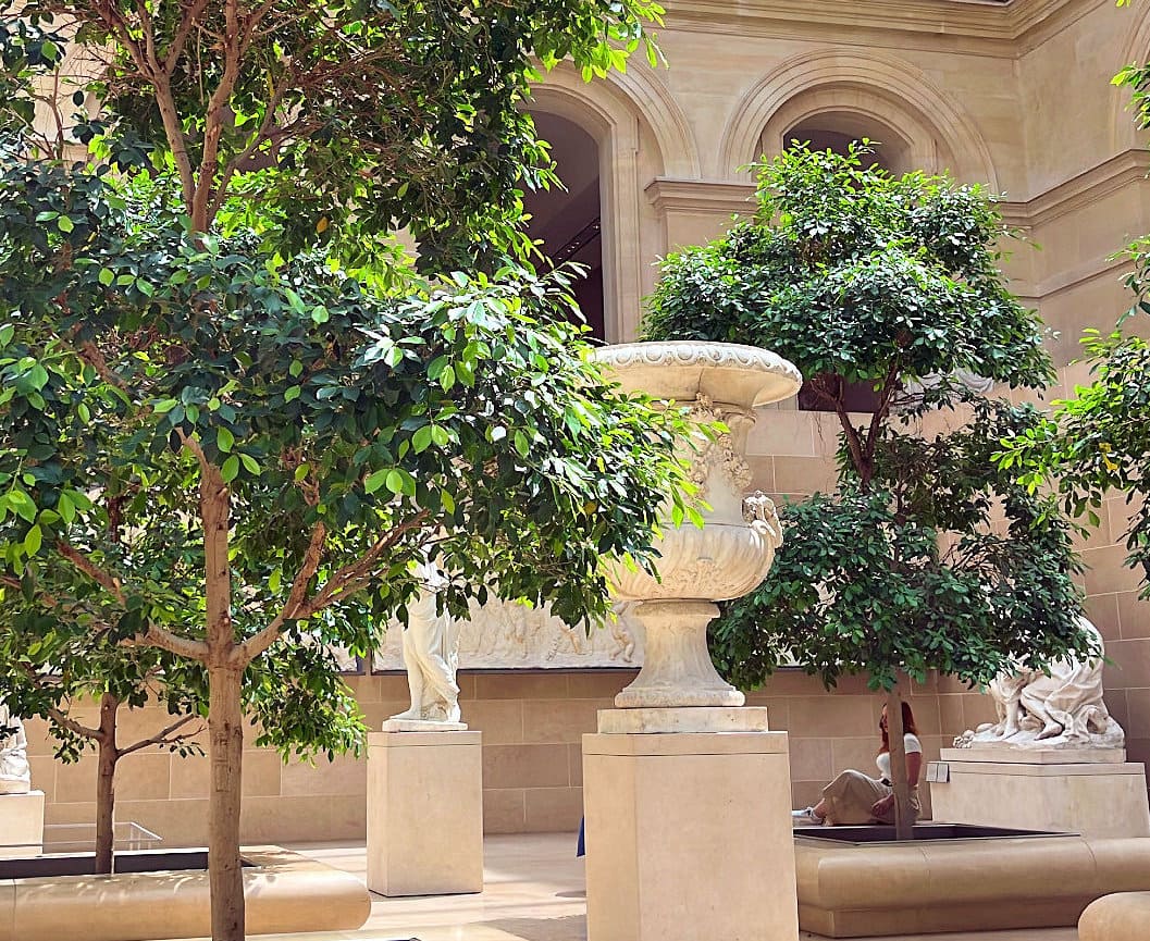 Sisäpiha, jossa on vihreitä puita, patsaita ja iso koristeellinen kivimalja, taustalla arkkitehtuuria ja istuva henkilö.