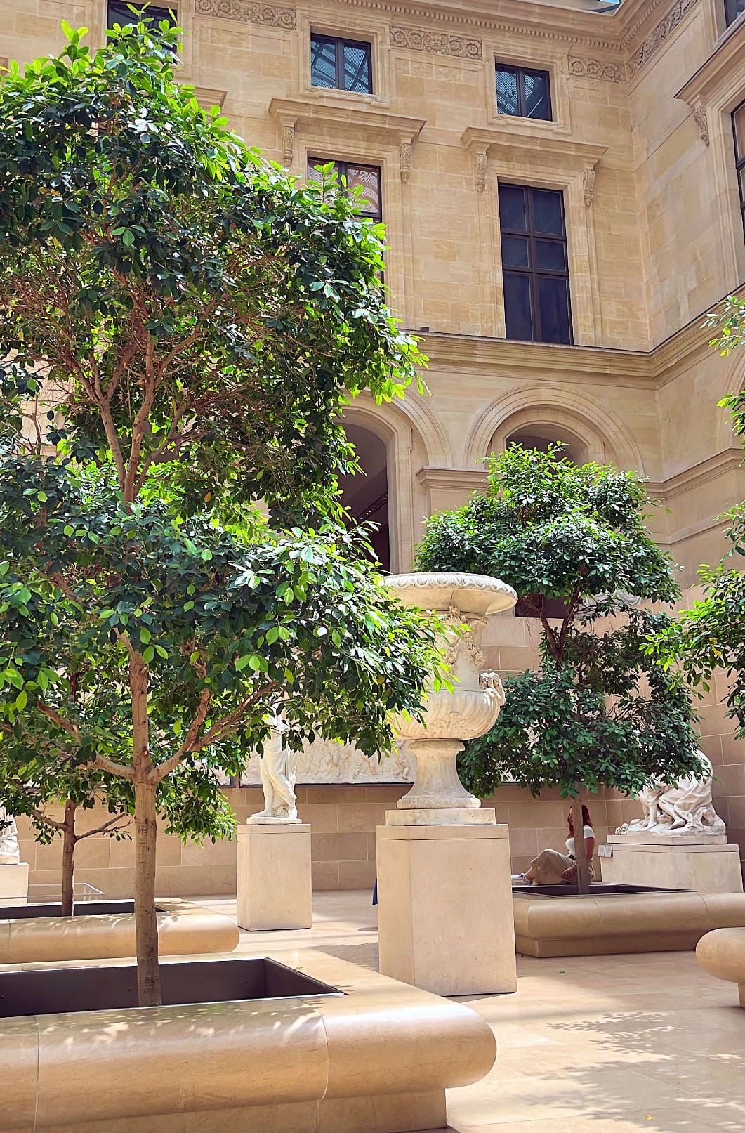 Valoisa museo-sisäpiha, jossa on vehreitä puita suurissa istutuslaatikossa, veistoksia ja koristeellinen samottimalja.