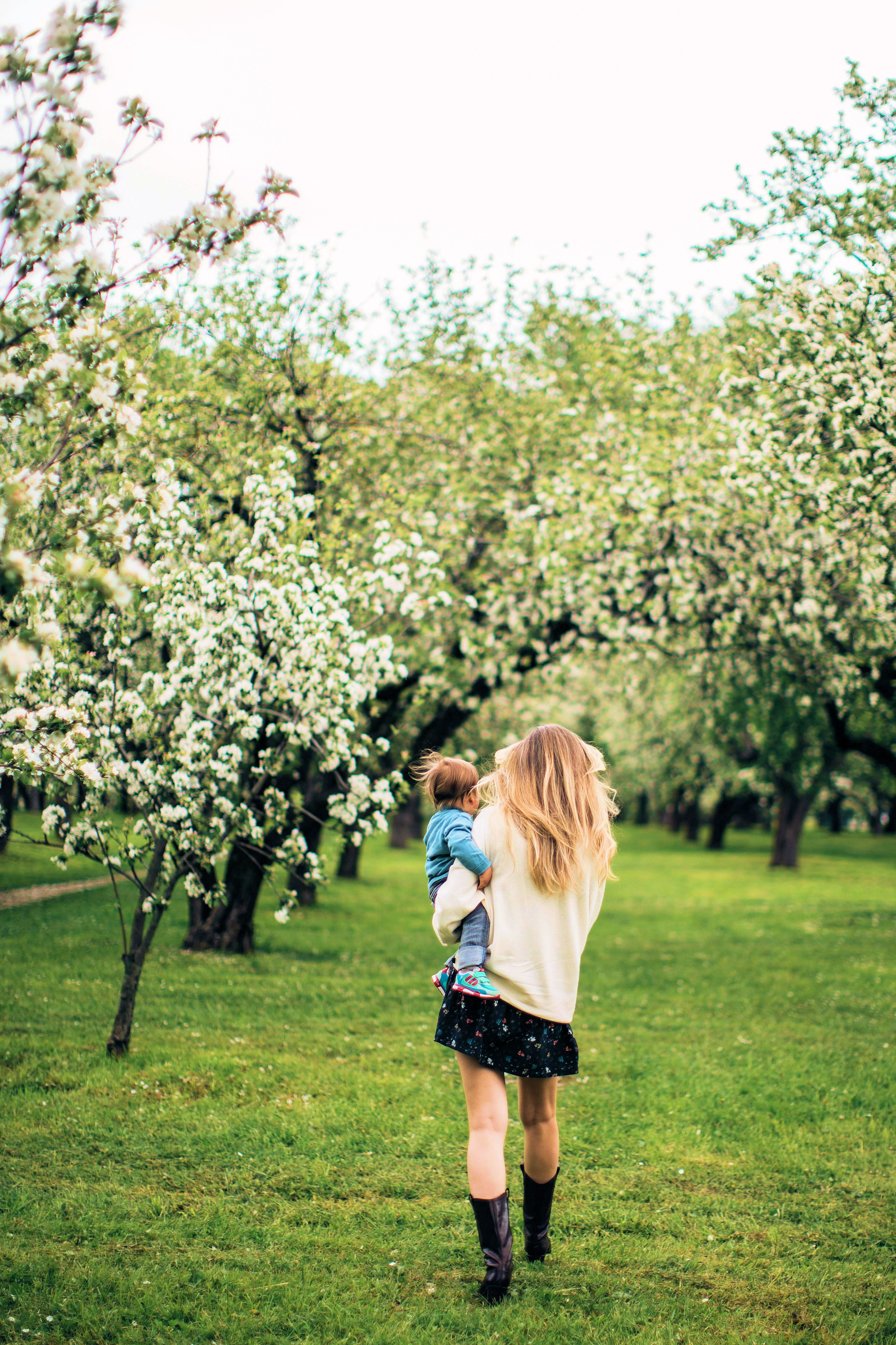 Nainen kävelee kukkivassa omenatarhassa kantaen lasta käsivarrellaan.