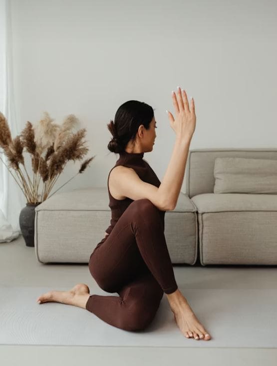 Nainen tekee joogavenytystä olohuoneessa harmaalla joogamatolla vaaleanharmaan sohvan edessä.