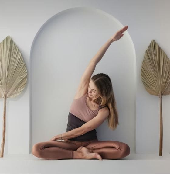 Nainen tekee sivutaivutusjoogaharjoituksen risti-istunnassa minimalistisessa tilassa, jossa on suurikokoinen kaareva seinä ja kaksi kuivattua lehteä.
