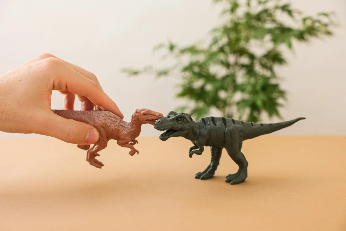 Kädessä ruskea dinosauruslelu toisessa pöydällä oleva musta dinosauruslelu, taustalla vihreä kasvi.